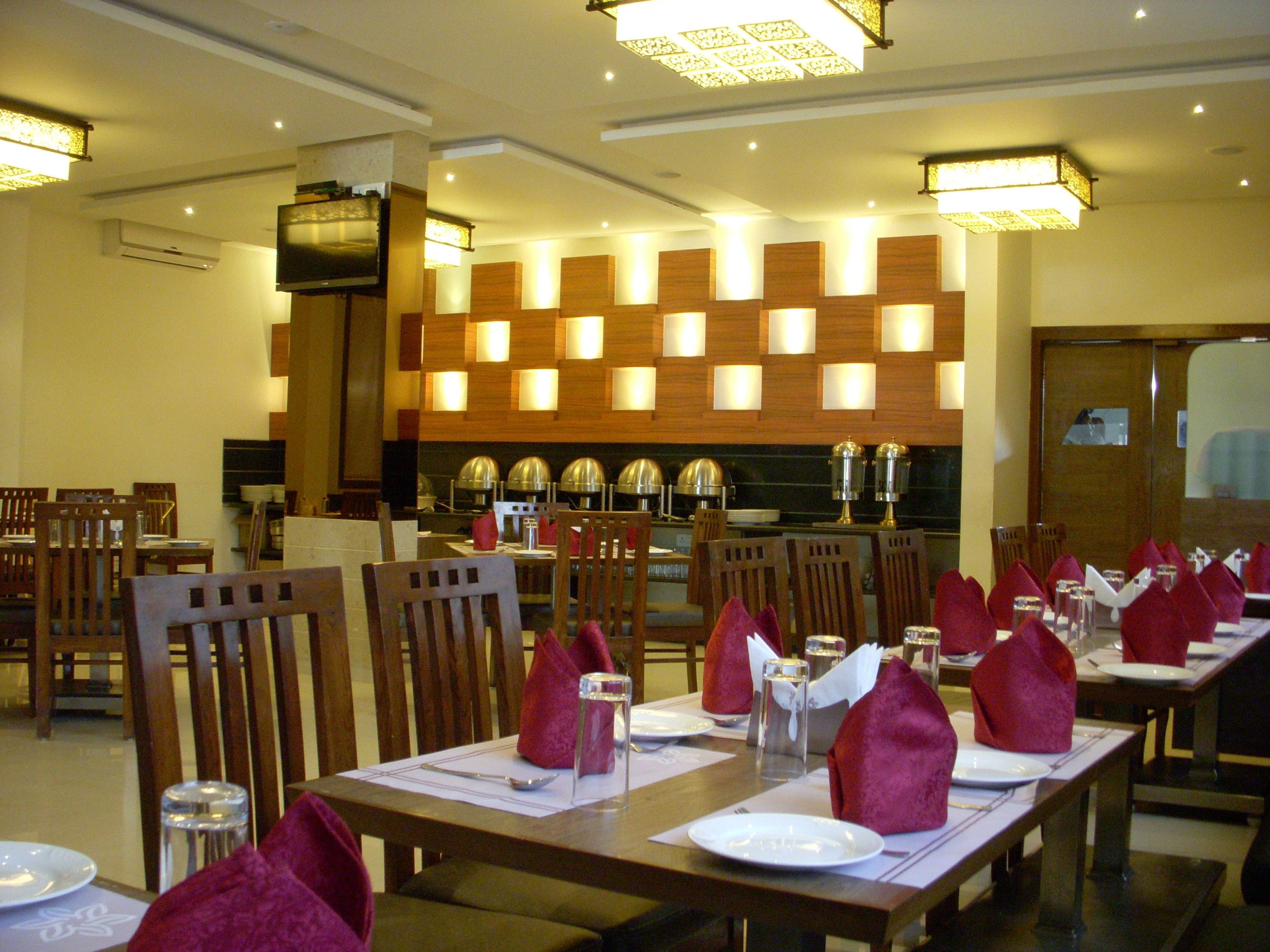A restaurant with multiple dining tables set with red napkins, glasses, and cutlery. There are wooden chairs and a kitchen area in the background.