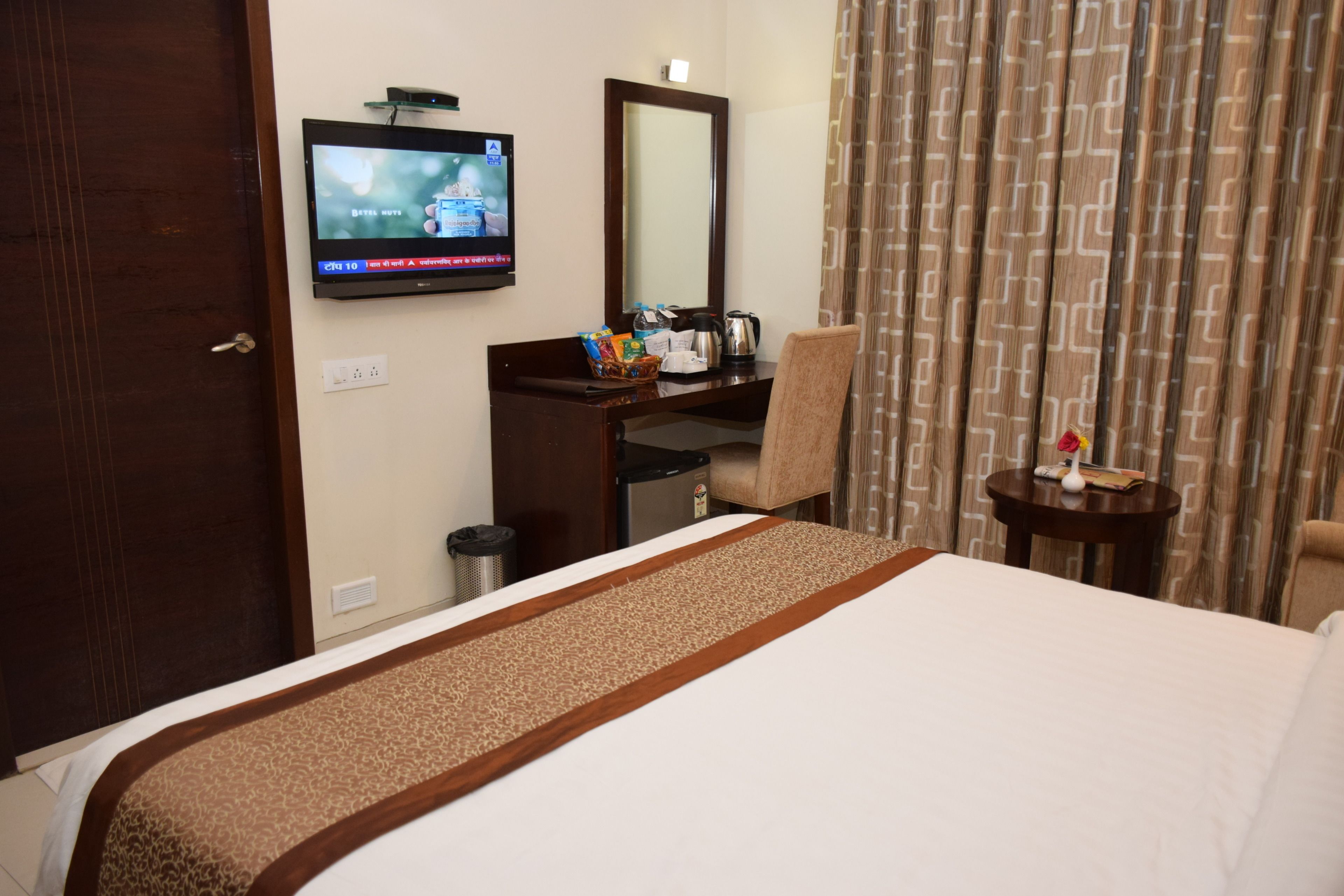 A hotel room with a bed, a desk with a chair, a television, a mirror, a small table with a vase, and curtains.