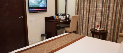 Habitación de hotel con cama, escritorio y silla, televisor, espejo, una mesita con un jarrón y cortinas.