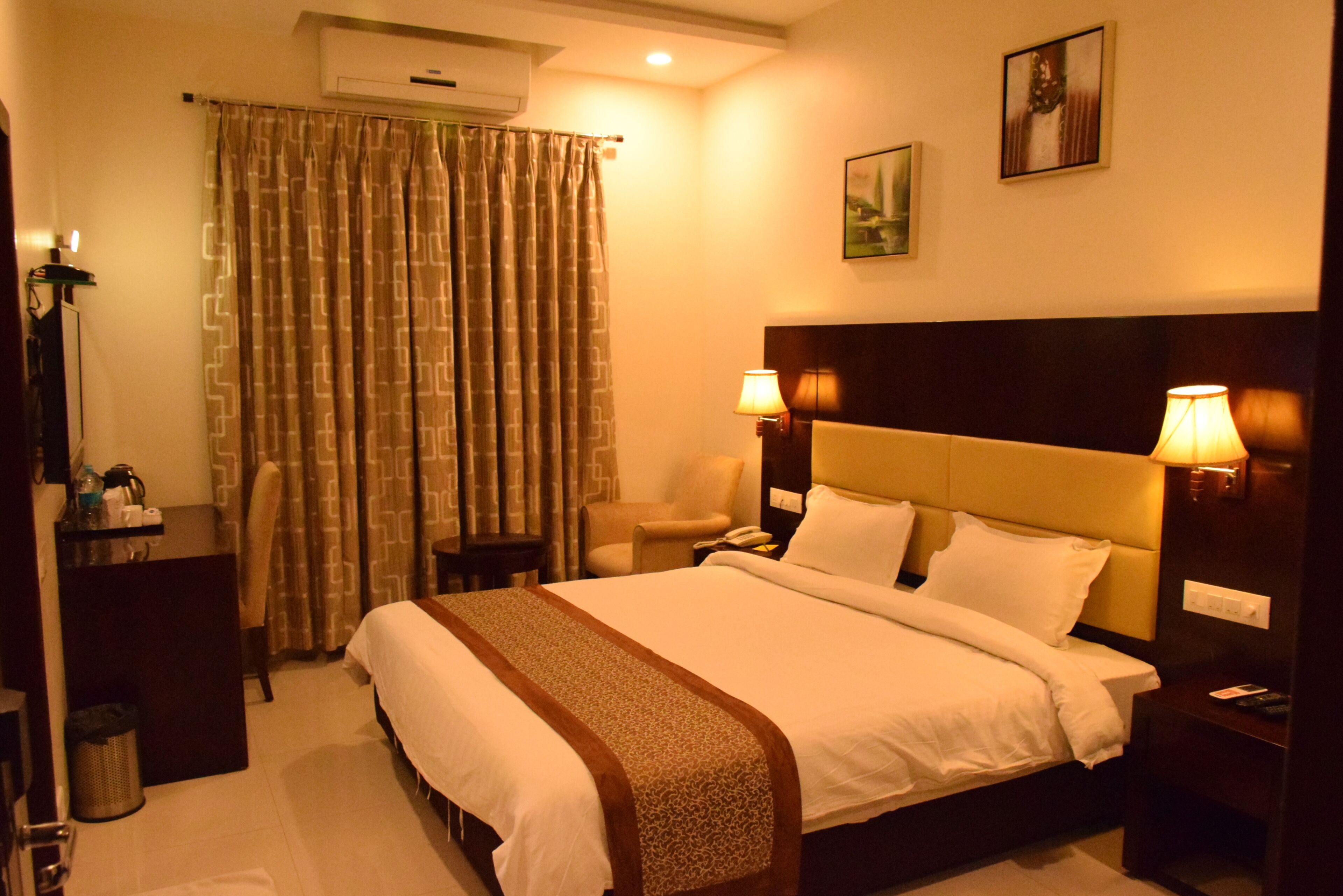 A hotel room with a bed, two bedside lamps, a chair, a desk, and an air conditioning unit.