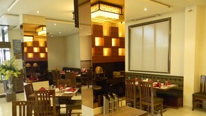 Um restaurante com mesas e cadeiras de madeira, um balcão central de atendimento e luminárias pendentes.