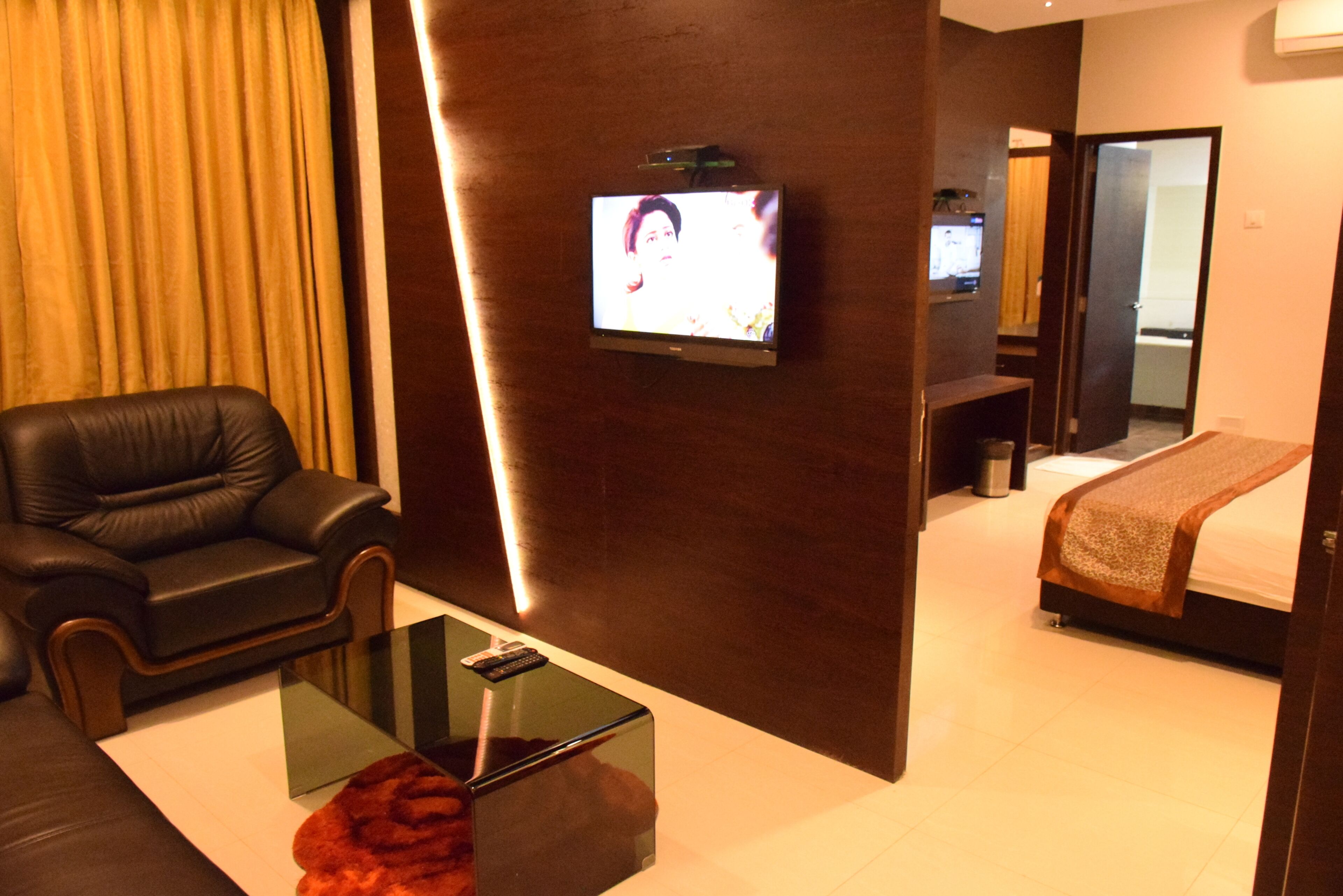 Una habitación de hotel con un televisor montado en la pared, un sillón de cuero, una mesa de centro de vidrio, una cama con una manta marrón y un espejo.