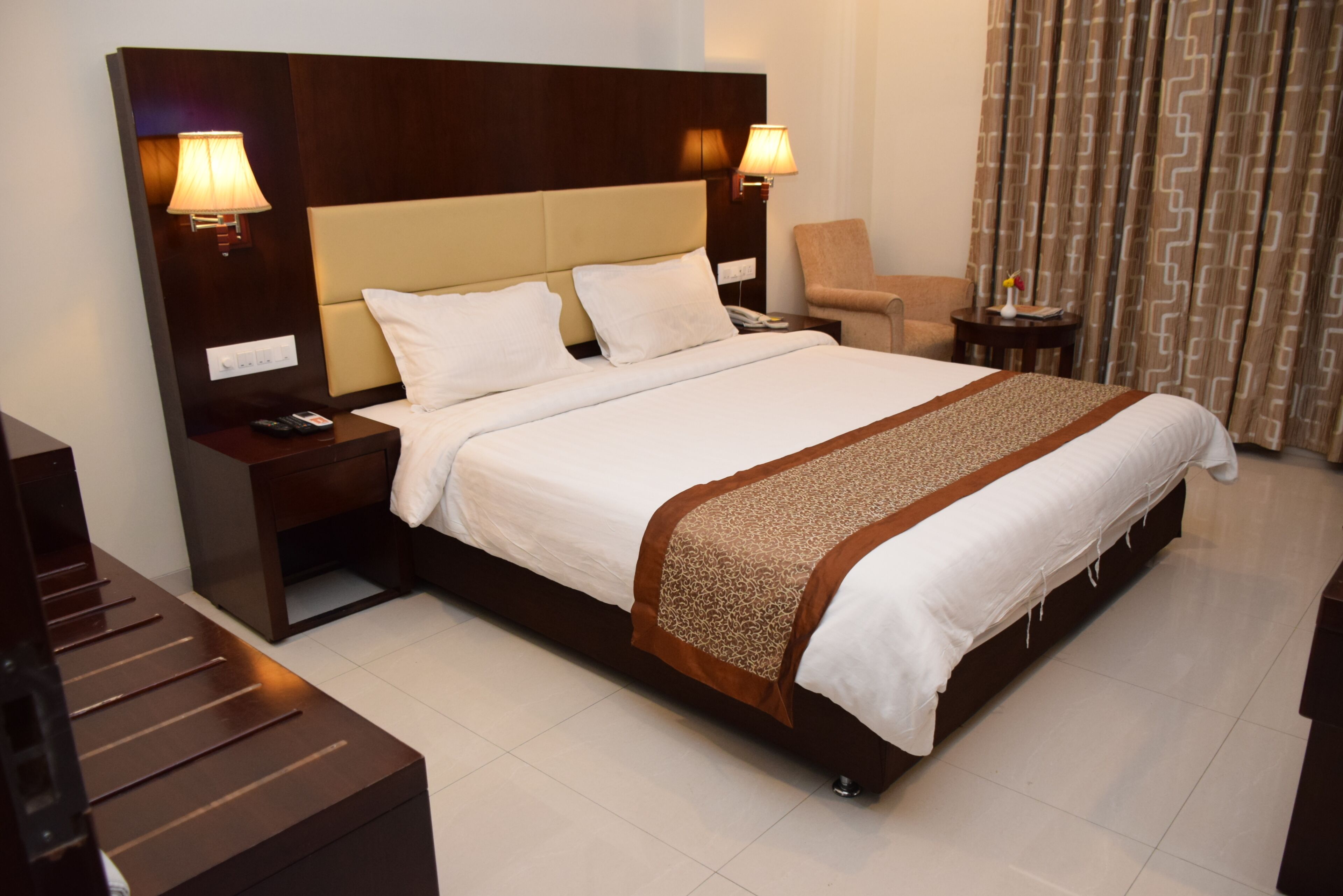 Una habitación de hotel con una cama grande, dos mesitas de noche con lámparas, una silla y una pequeña mesa con un florero.