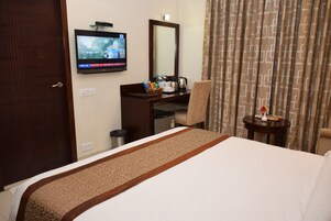 Habitación de hotel con cama, escritorio con televisor, silla, mesita con un florero y cortinas.