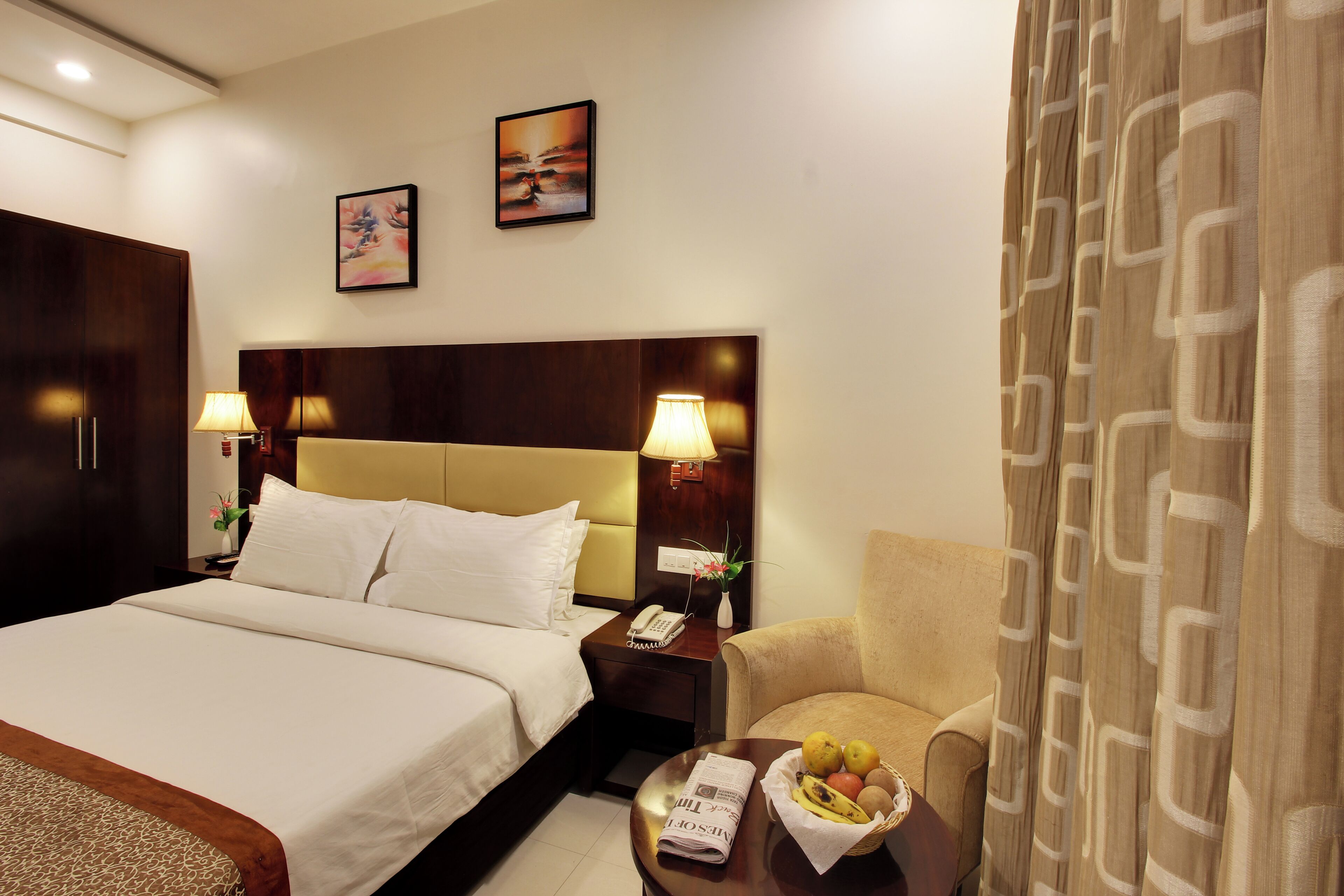 A hotel room with a large bed, bedside lamps, a nightstand with a phone, a chair, and a table with fruit.
