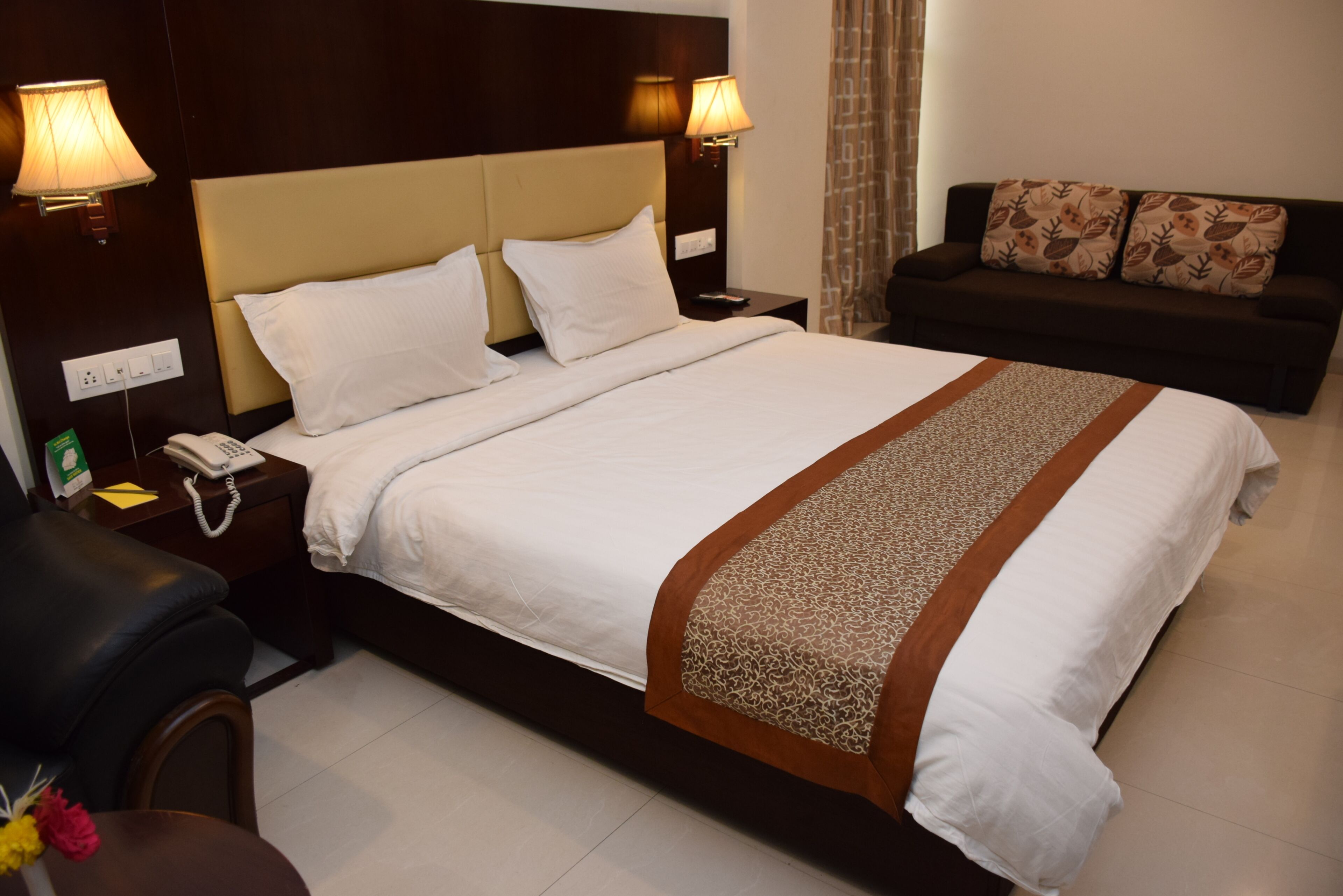 A hotel room with a large bed, two bedside lamps, a bedside phone, a sofa with patterned cushions, and a small table with a vase of flowers.