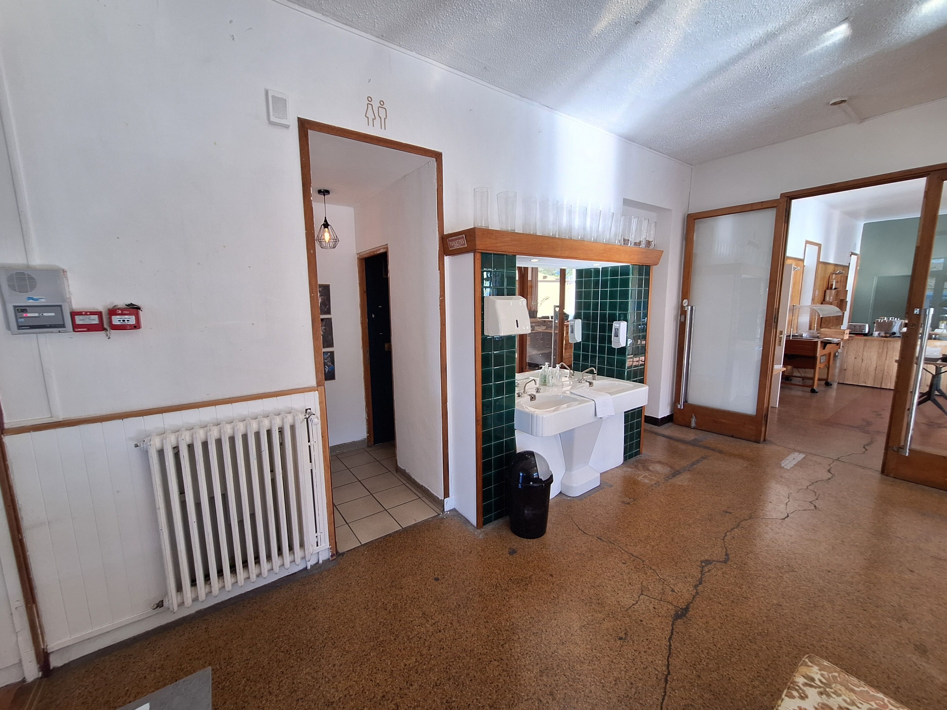 Un couloir d’hôtel avec un radiateur, une poubelle, un lavabo avec une serviette et une porte donnant sur une chambre.