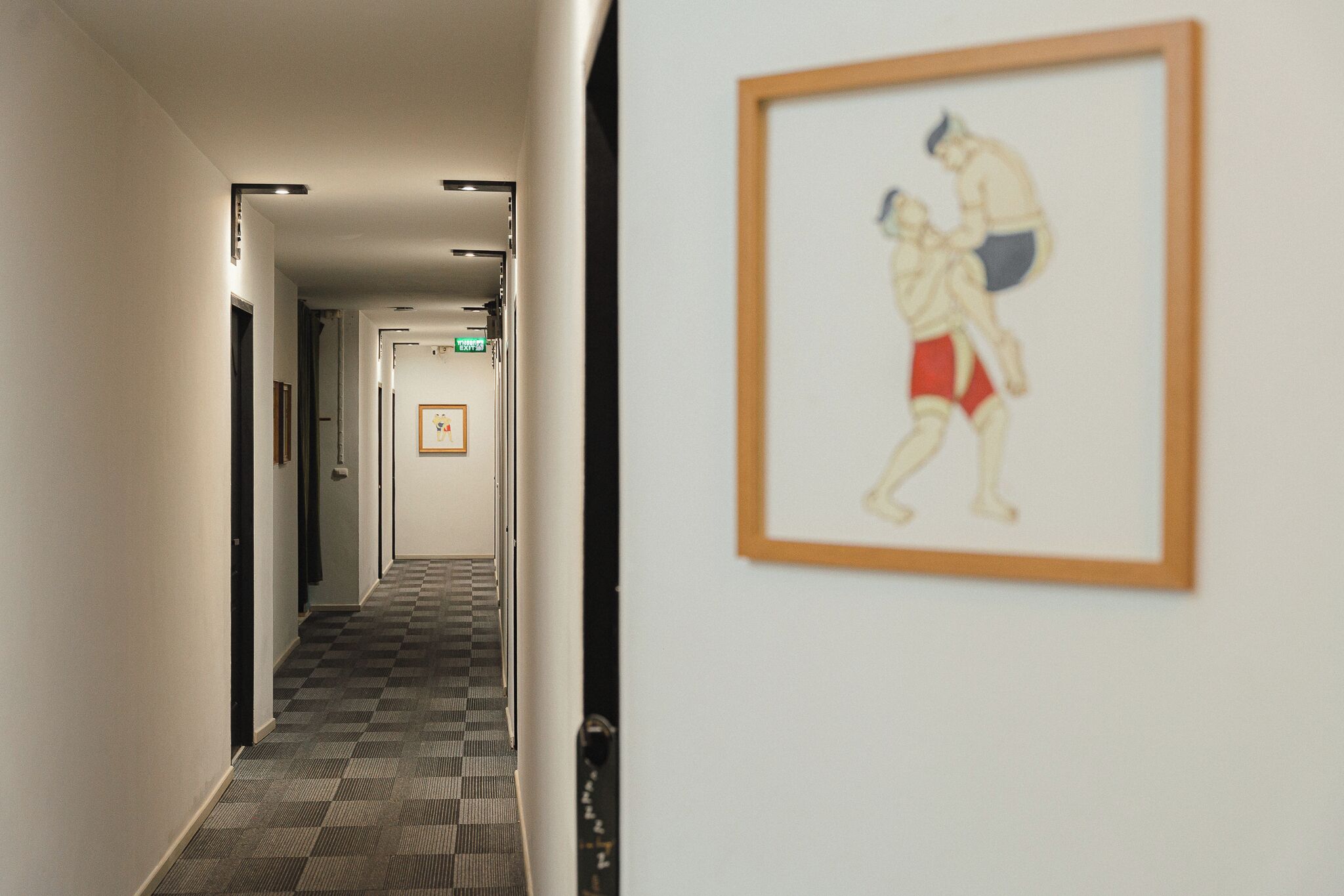 A hotel hallway with a checkered floor, framed artwork on the wall, and doors leading to rooms.