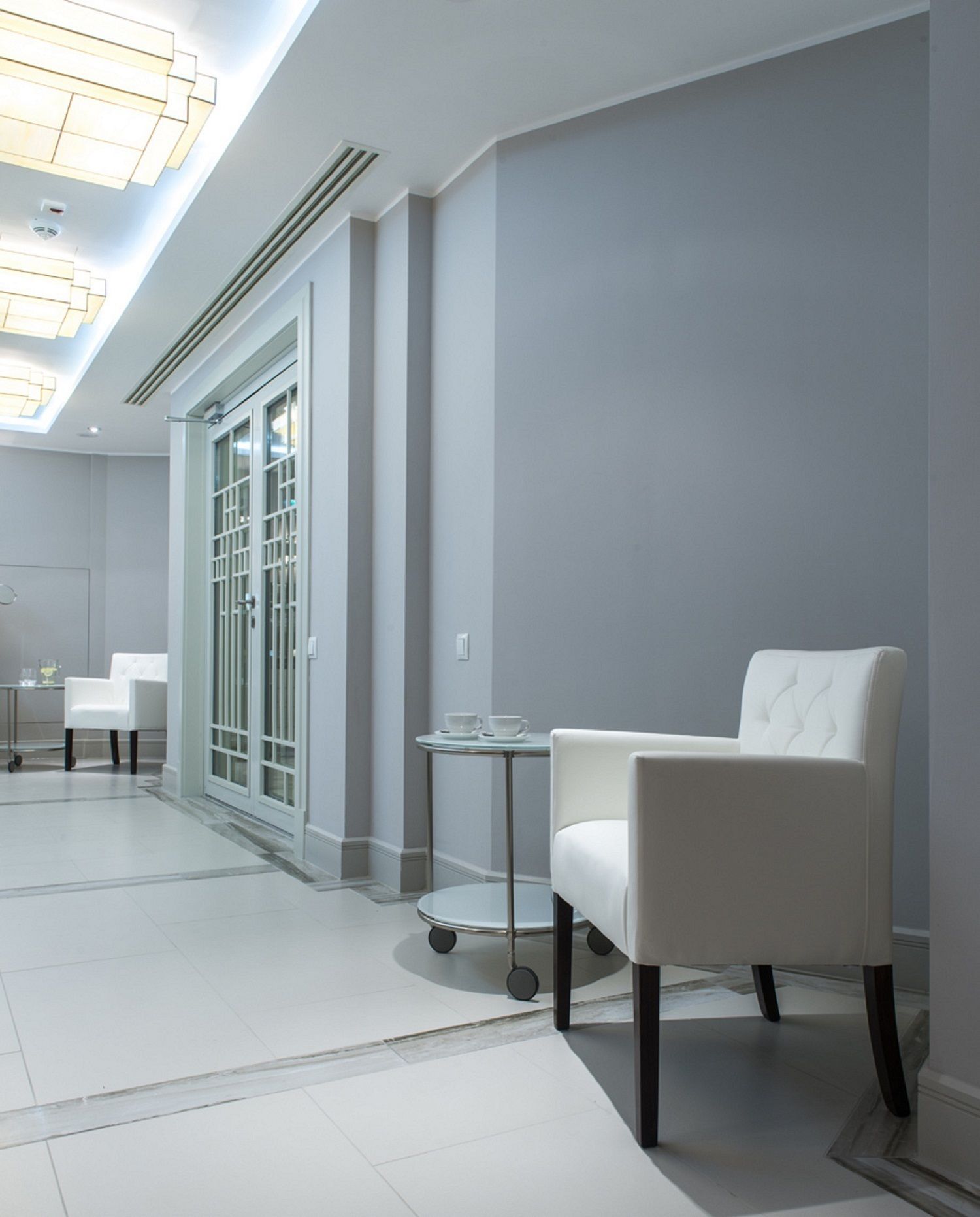 A modern hotel lobby with a white chair, a small round table, and a glass door leading to a balcony.