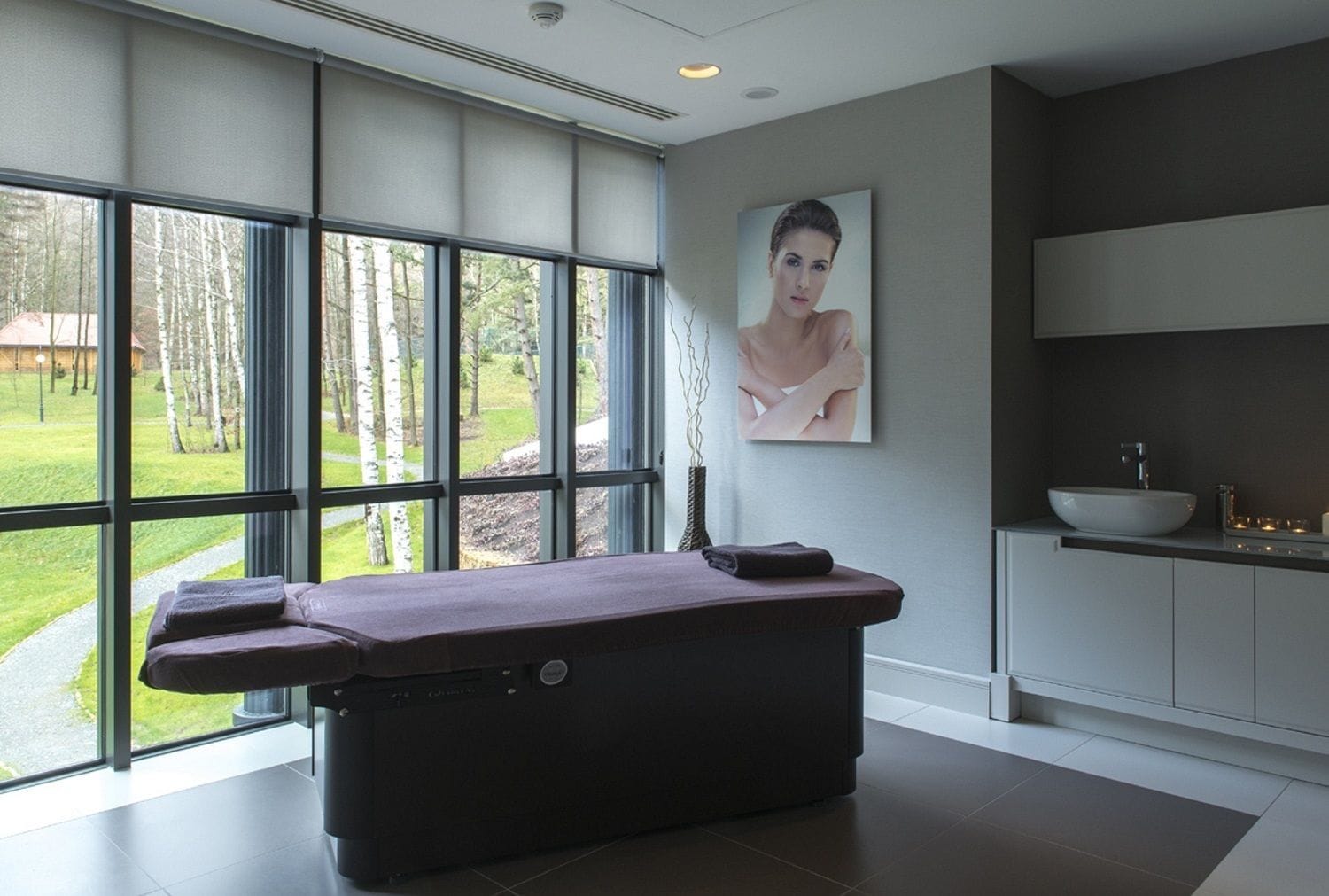 Un moderno spa con camilla de masaje, ventanas amplias, un lavabo y un cuadro.