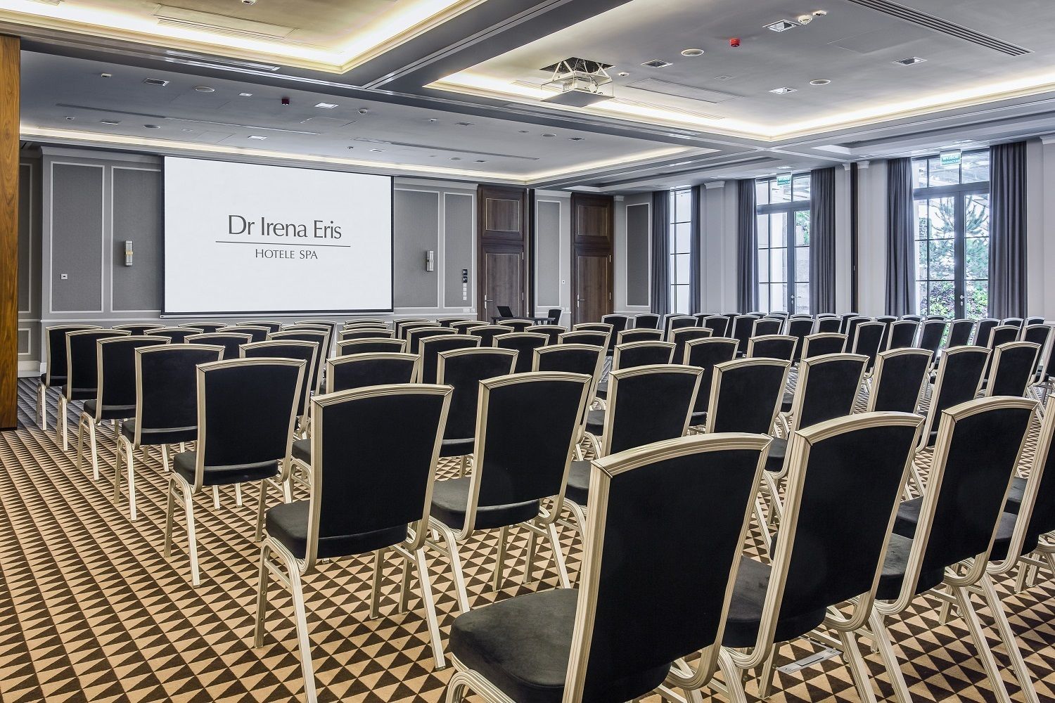 A conference room with rows of black chairs, a projector screen displaying 'Dr Helena Elis HOTELE SPA', and large windows with curtains.