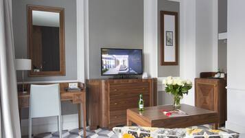 A modern hotel room with a wooden desk, a television, a dining table with a vase of flowers, and a patterned sofa.