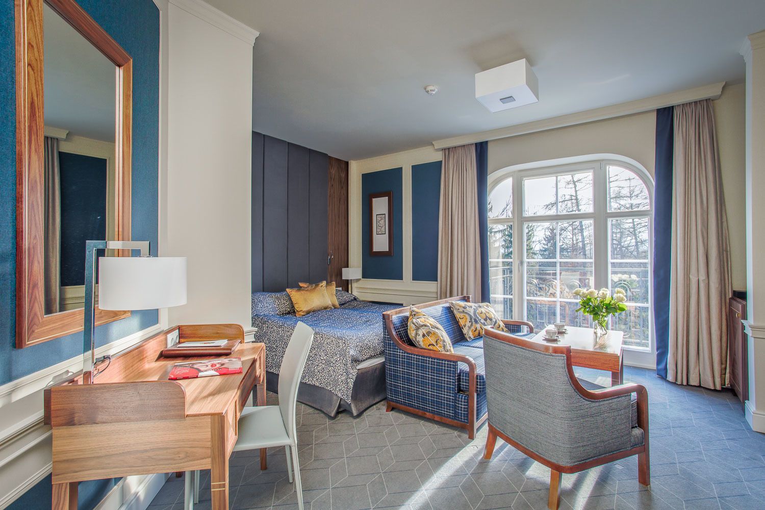 A hotel room with a bed, a desk, a chair, a window with curtains, and a view of trees.