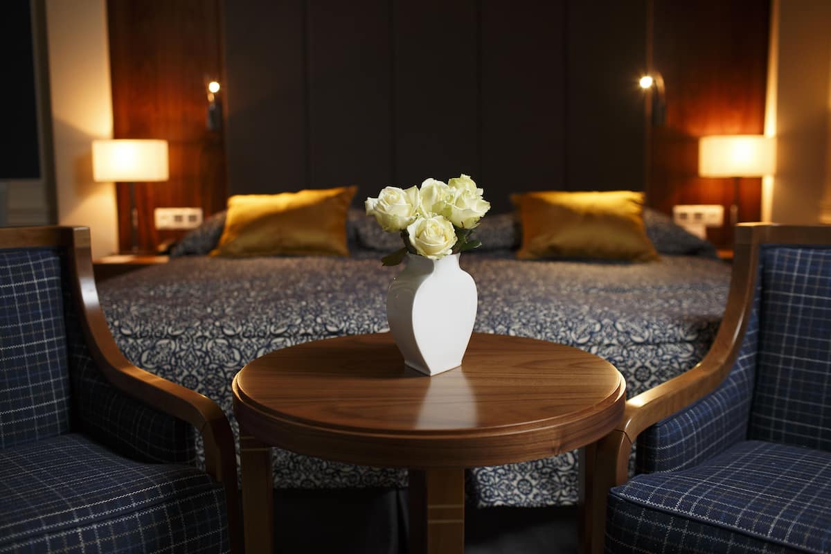 Une chambre d’hôtel comprenant un lit, deux lampes de chevet, une table ronde en bois ornée d’un vase de fleurs et deux fauteuils rembourrés.