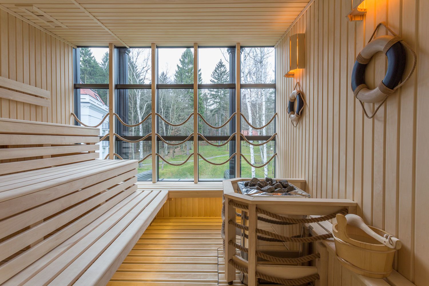 A sauna room with wooden benches, a wooden table with a tray of stones, and a view of trees through large windows.