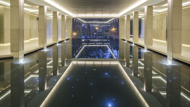 A modern hotel corridor with reflective floors, glass walls, and a central view of a cityscape at night.