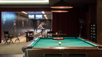Ein Billardtisch mit Spielball und bunten Kugeln, eine Bowlingbahn und ein großer Bildschirm, der ein Sportereignis zeigt.