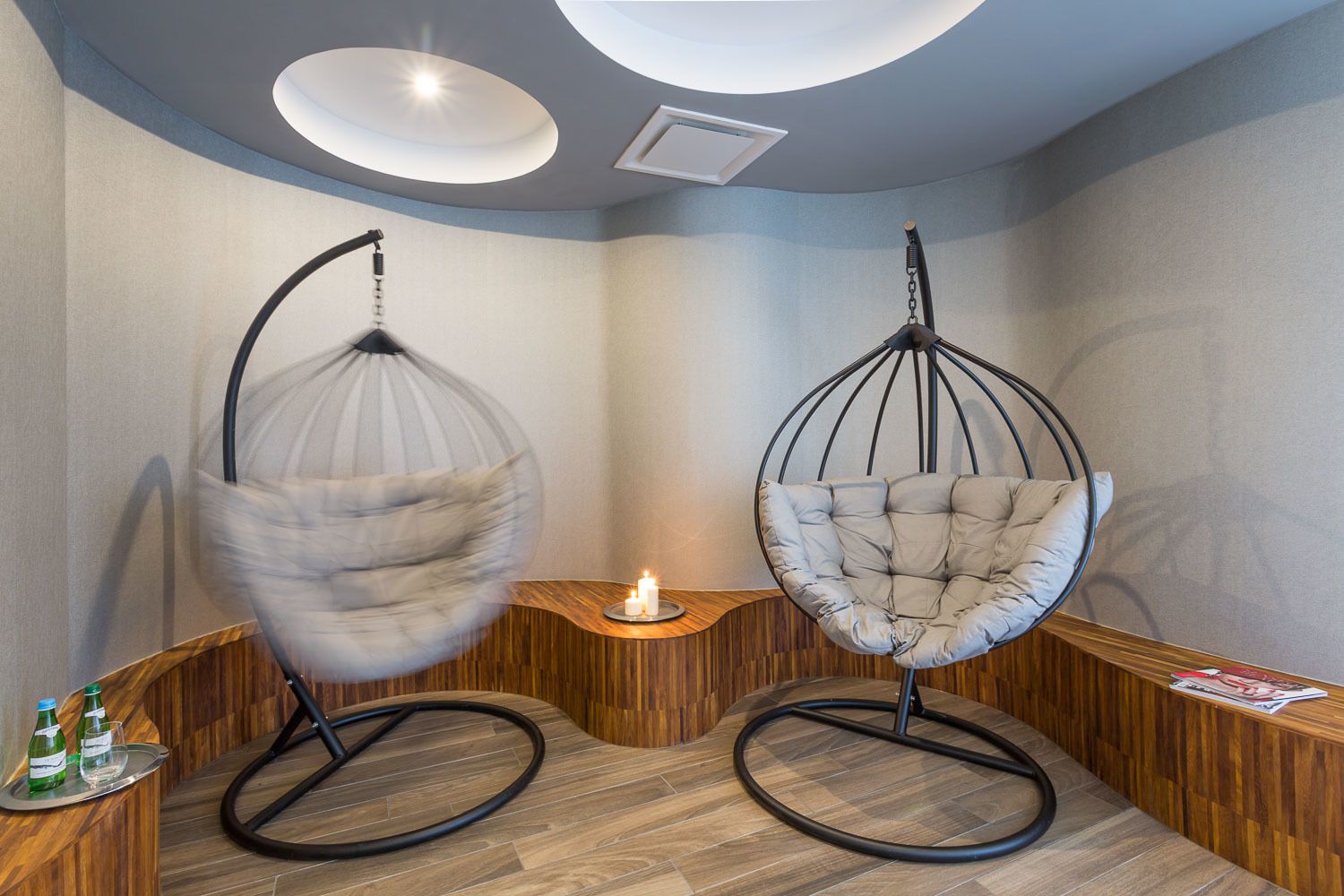 Dos sillas colgantes en una habitación con suelo de madera, una mesita con una vela y una revista sobre un mostrador de madera.