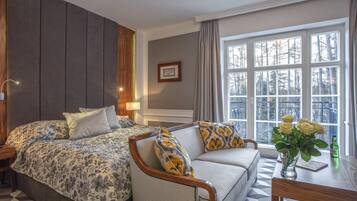 Ein Hotelzimmer mit einem großen Bett, einer Couch, einem Tisch mit Blumenvase und einem Fenster mit Blick auf Bäume.