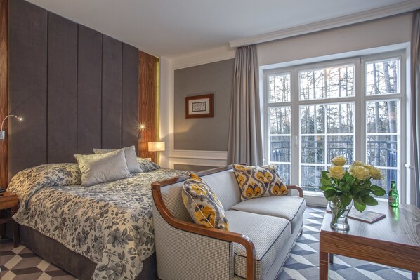 A hotel room with a large bed, a sofa, a table with a vase of flowers, and a window with a view of trees.