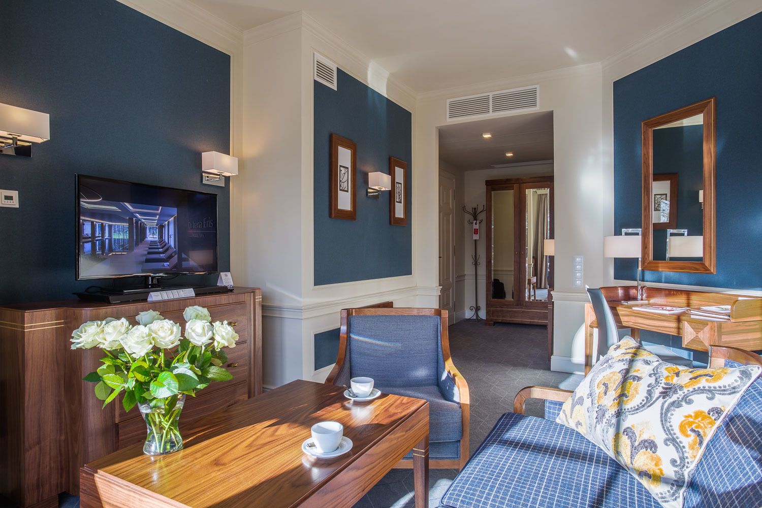A hotel room with a TV, a wooden desk with a vase of flowers, a sofa, and a bed with a patterned bedspread.