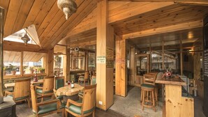 A wooden interior with round tables and green cushioned chairs, a bar area, and large windows allowing natural light. - Whispering Inn by Vivaan (Manali)