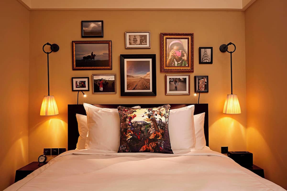 A hotel room with a bed, two bedside lamps, a nightstand with a clock, and a wall decorated with framed pictures.