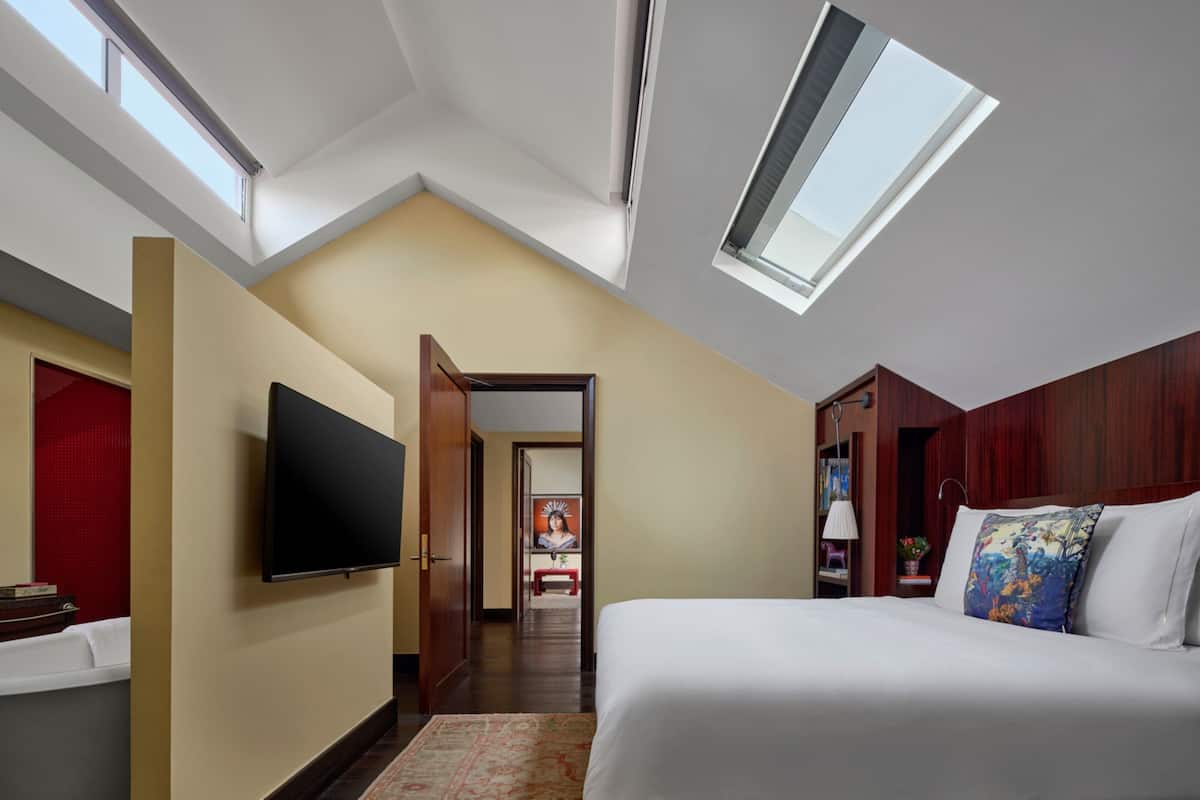 A modern hotel room with a sloped ceiling, skylight, a flat-screen TV, a bed with white linens, and a wooden wardrobe.