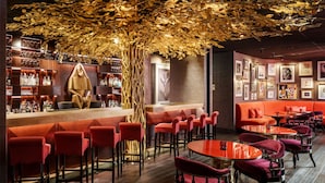 A bar area with a wooden bar counter, red bar stools, and a decorative tree overhead. There are framed pictures on the walls and a red sofa in the seating area. - The Vagabond Club, Singapore, a Tribute Portfolio Hotel (Singapore)