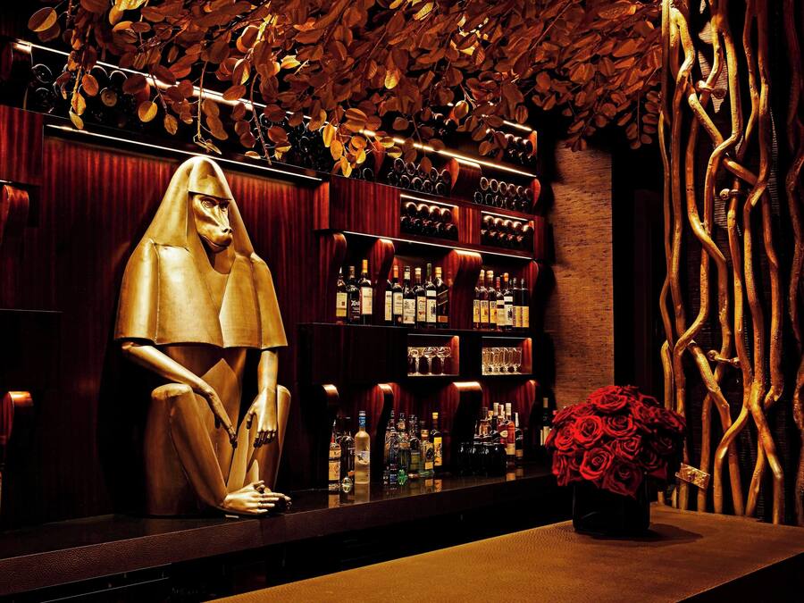 Un bar con un maniquí vestido con una túnica dorada, estantes llenos de botellas de licor y un jarrón de rosas rojas.