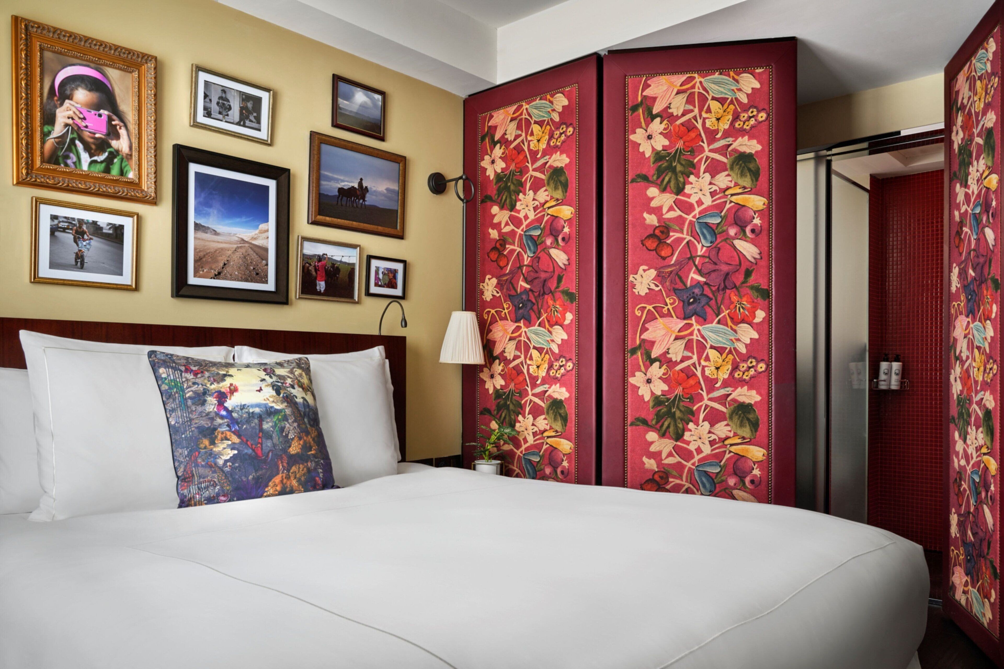 A hotel room with a large bed, multiple framed pictures on the wall, a floral-patterned partition, and a red door.