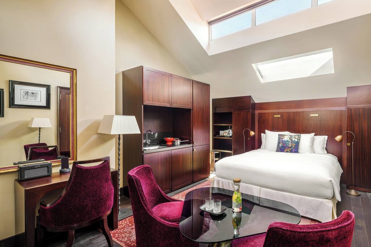 A hotel room with a bed, a desk with a chair, a small table with a glass top, a lamp, and a large window.