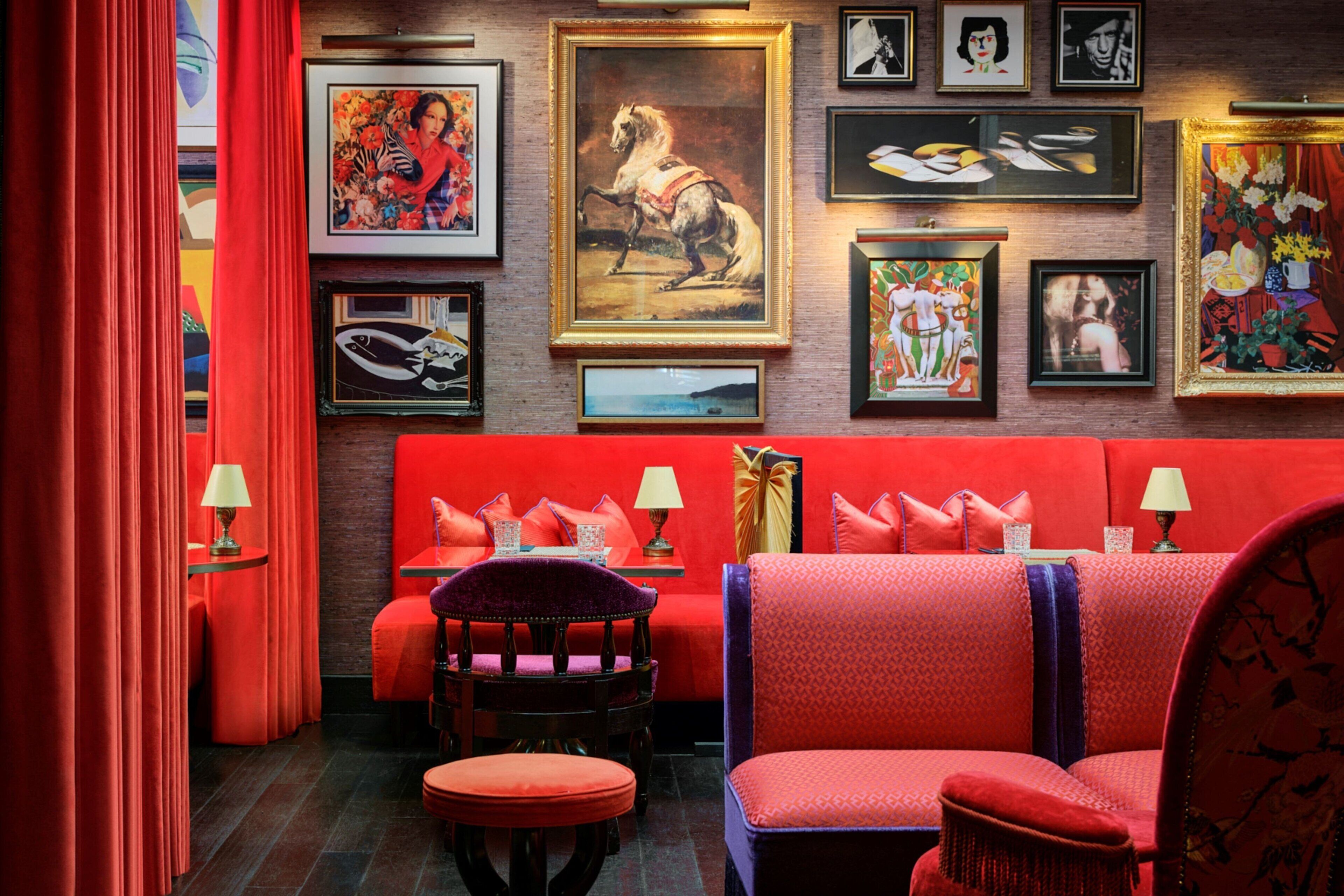 Habitación vibrante con paredes rojas, un sofá y una silla. Se observan obras de arte enmarcadas en la pared y una lámpara sobre la mesita de noche.