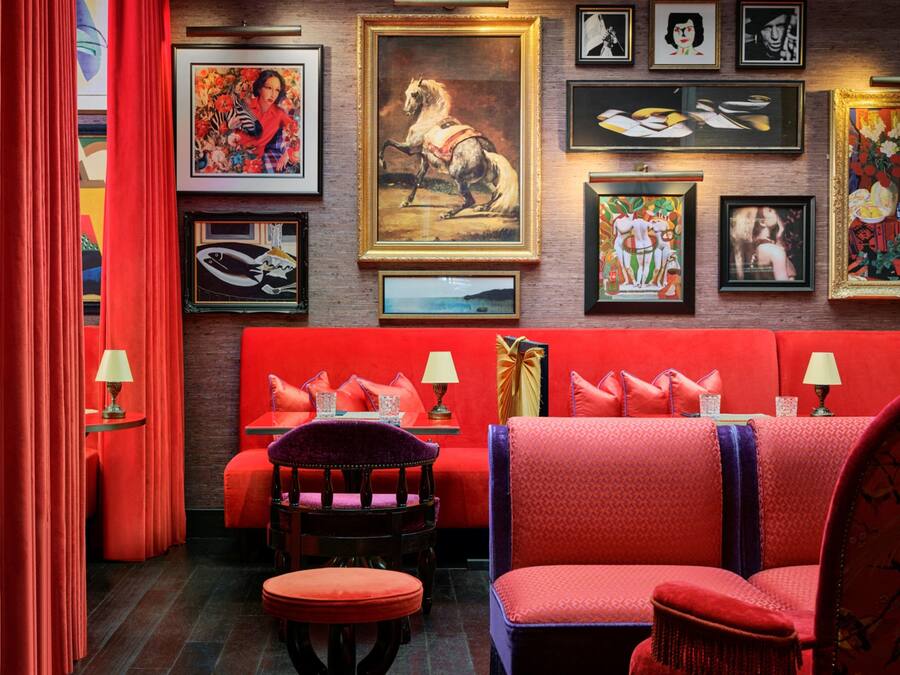 Habitación vibrante con paredes rojas, un sofá y una silla. Se observan obras de arte enmarcadas en la pared y una lámpara sobre la mesita de noche.