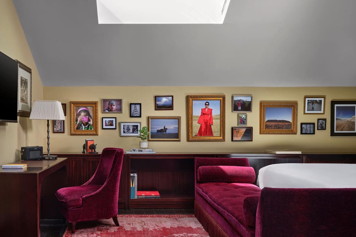 A hotel room with a bed, a desk with a lamp, a red armchair, a sofa, and a skylight.