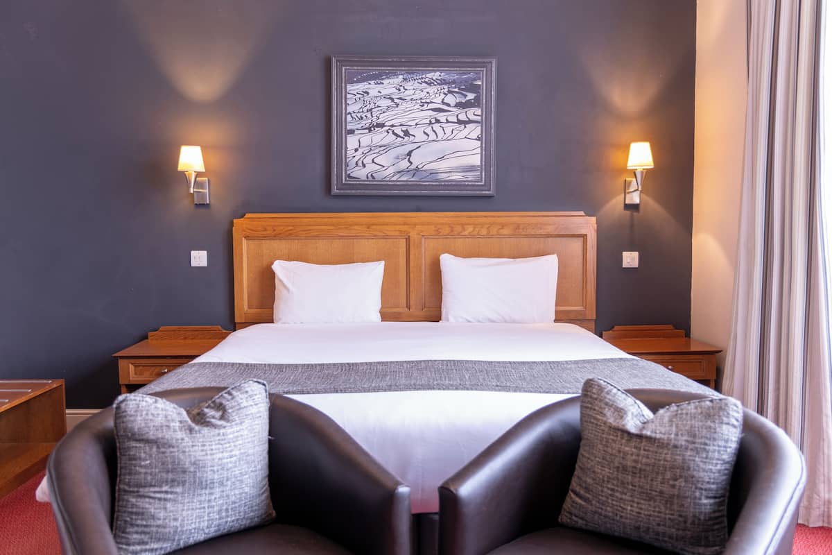 Habitación de hotel con una cama grande, dos mesitas de noche, cabecera, una obra de arte colgada en la pared y dos apliques, además de vistas al exterior a través de una ventana.