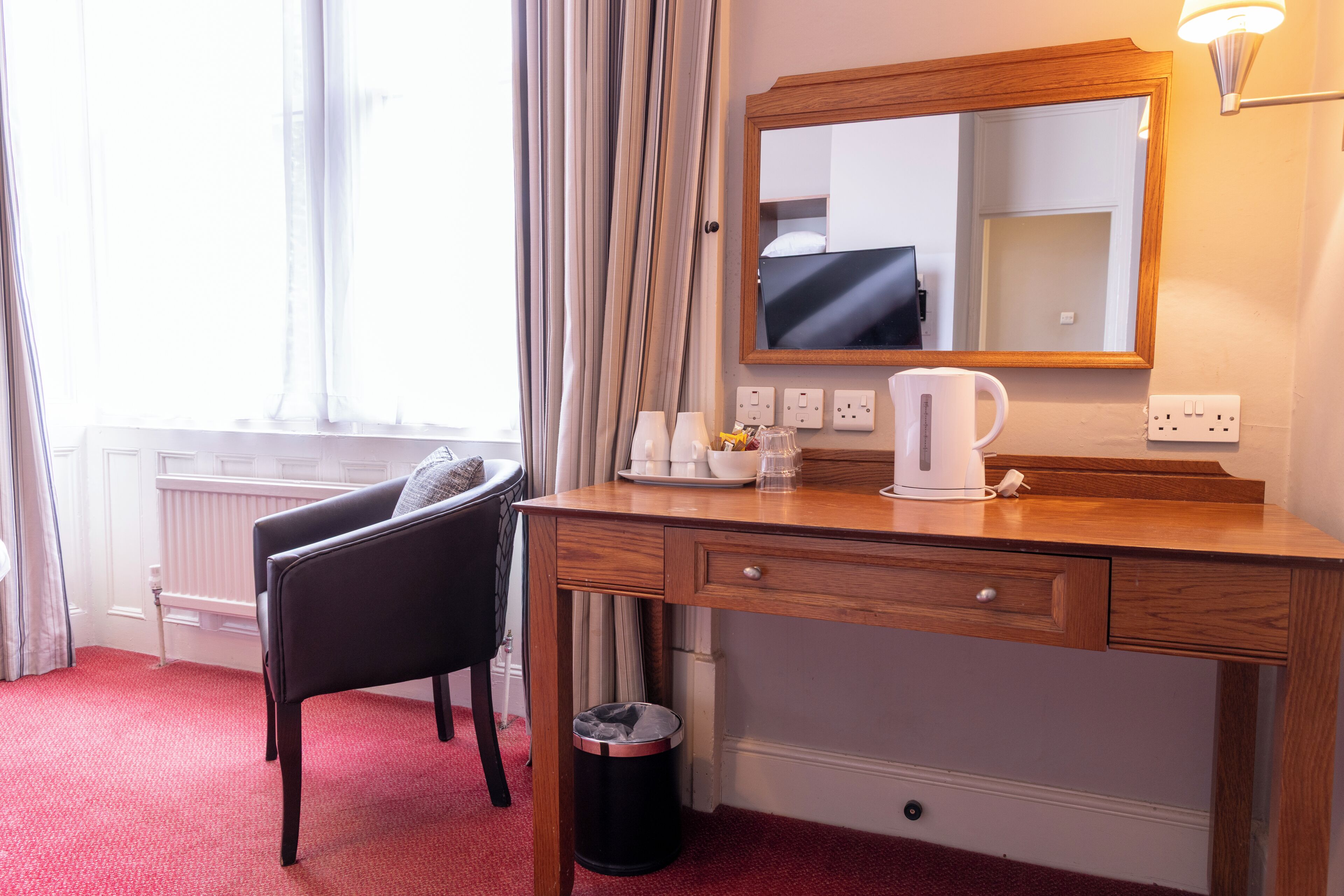 A hotel room with a wooden desk, a chair, a mirror, a coffee maker, and a radiator.
