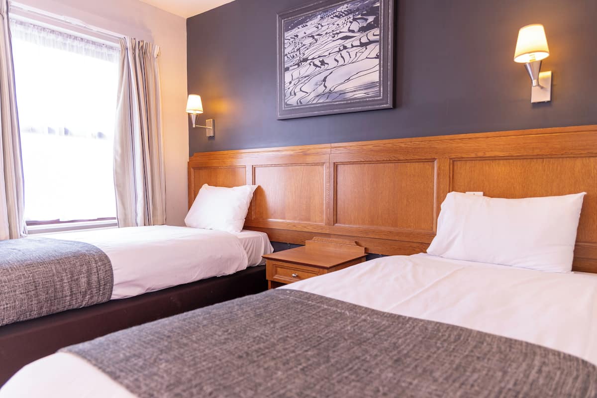 Una habitación de hotel con dos camas, cabecera de madera, mesita de noche, lámpara, ventana con cortinas e imagen enmarcada en la pared.