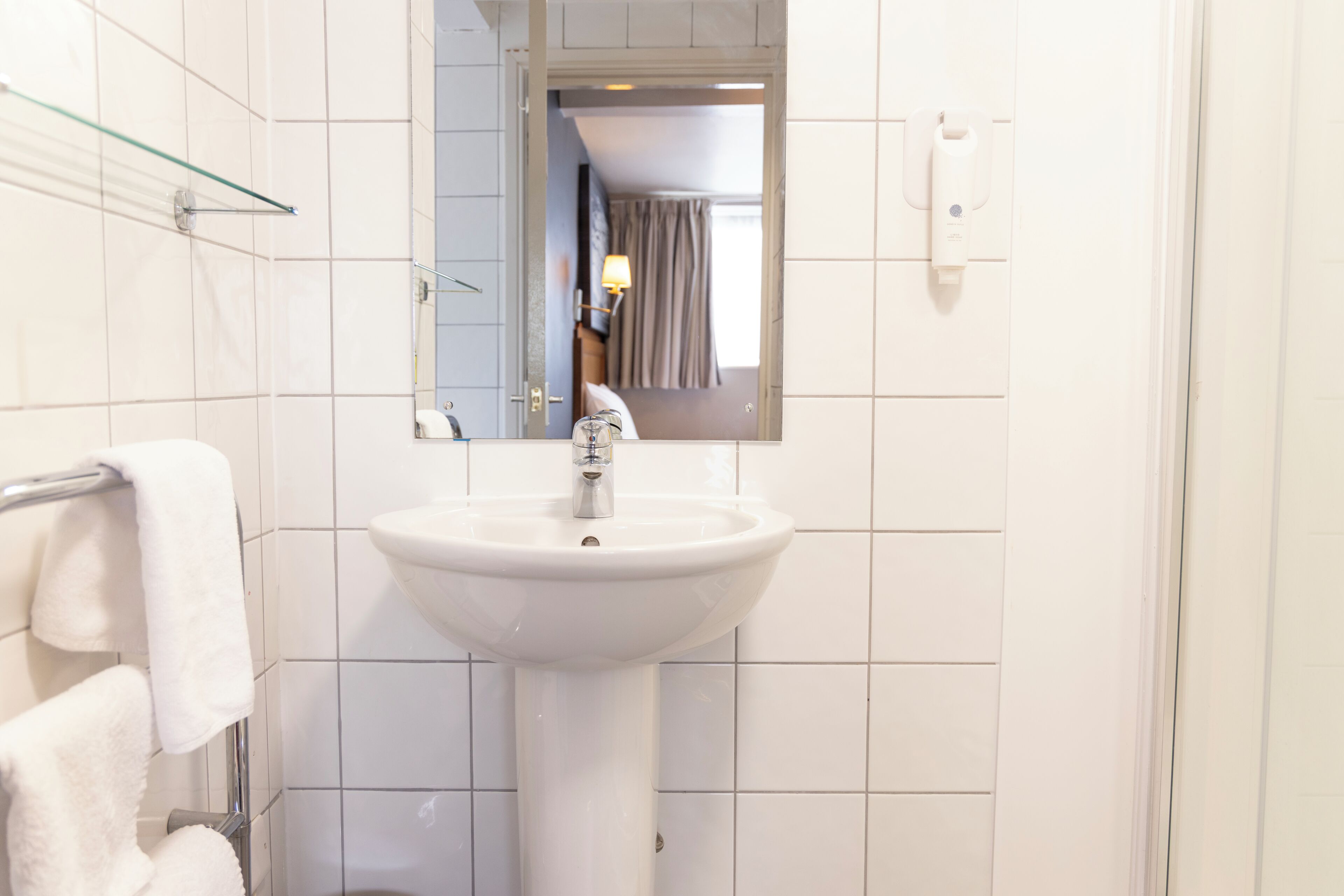 Un baño con un lavamanos blanco, un estante de vidrio y un espejo que refleja una habitación con una cama y cortinas.
