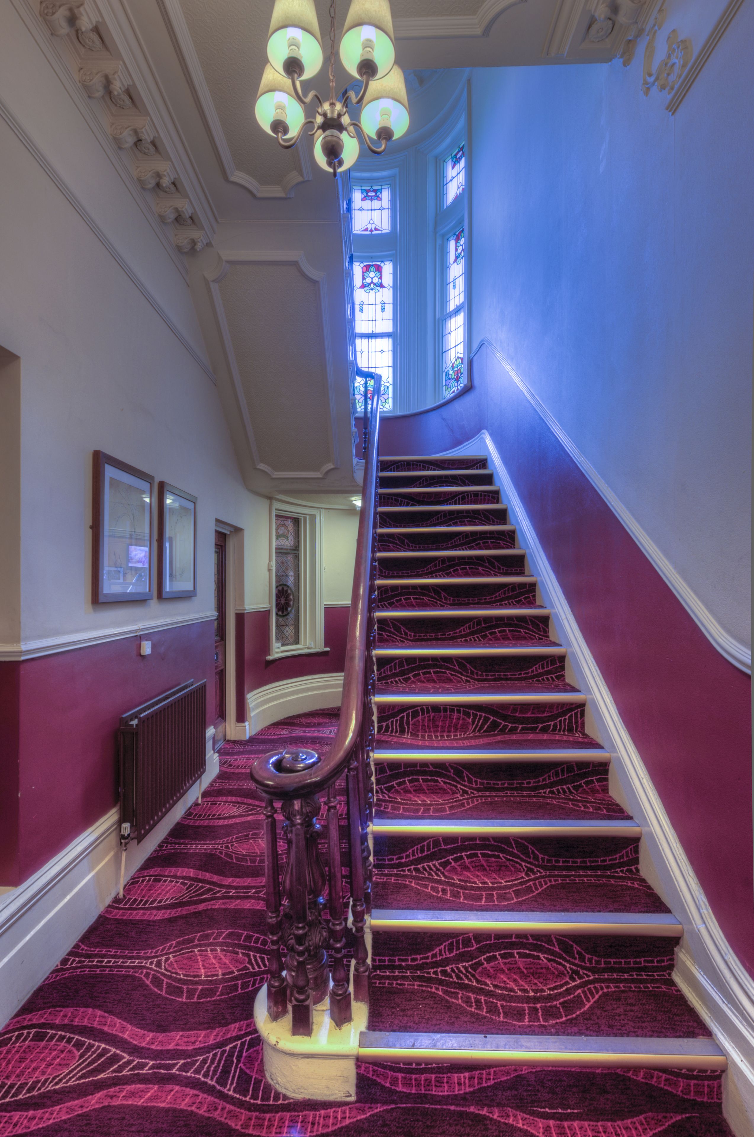 A grand staircase with ornate railings and a patterned carpet, leading to a room with a large window and a radiator on the wall.