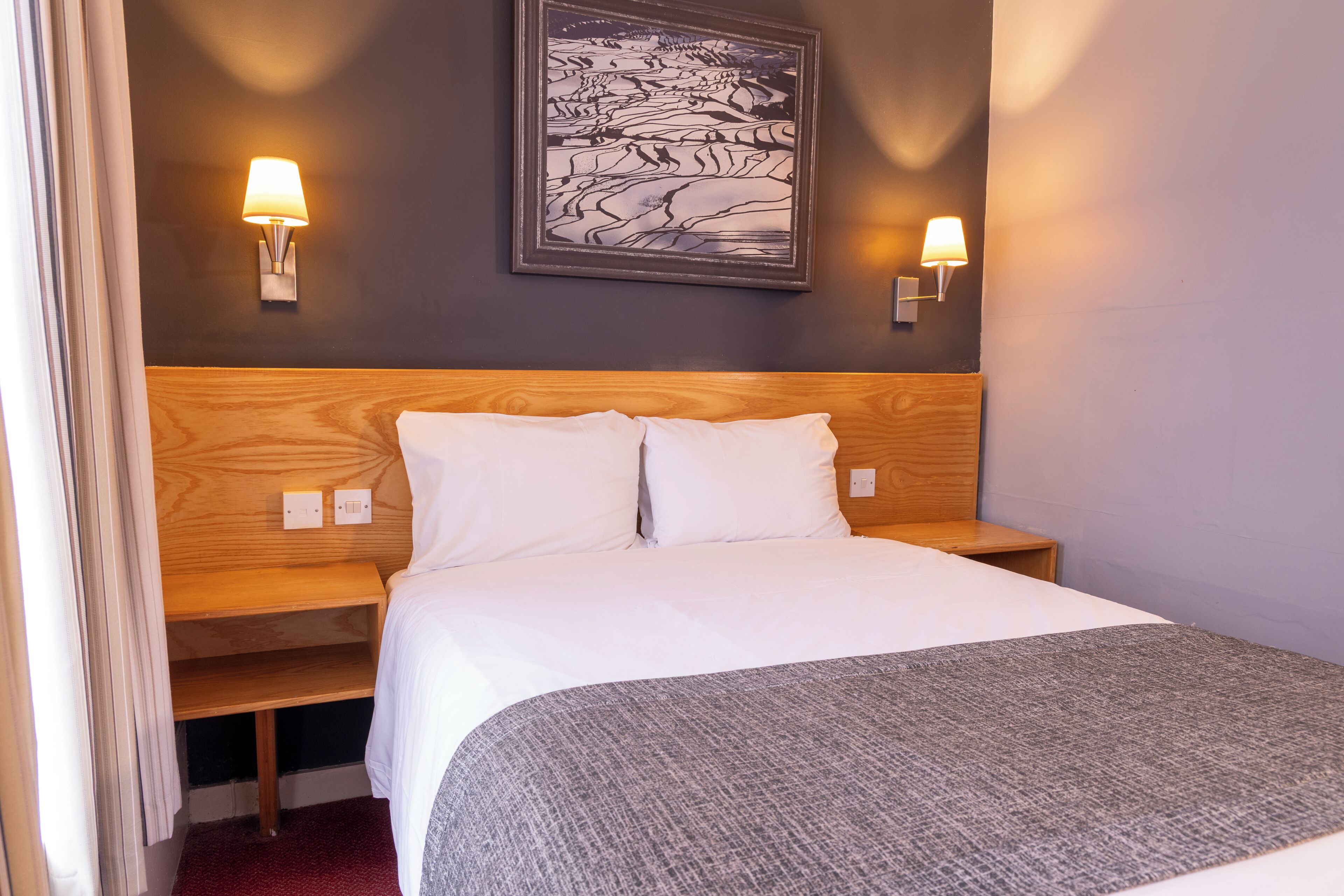 A hotel room with a wooden headboard, a large bed with white bedding, two bedside lamps, and a framed artwork on the wall.