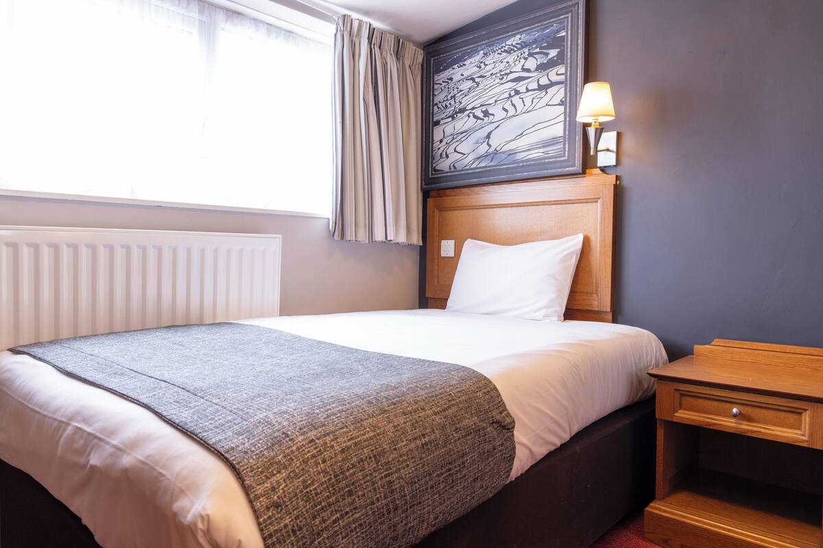 Una habitación de hotel con cama, mesita de noche, lámpara, ventana con cortinas y un cuadro en la pared.