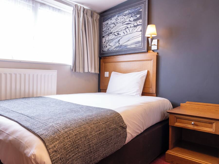 A hotel room with a bed, a nightstand, a lamp, a window with curtains, and a painting on the wall.