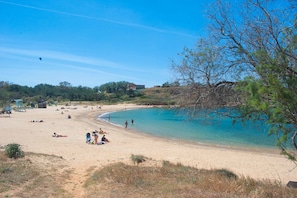 En strand med klart turkis vand, sandet bredde og sparsom vegetation.