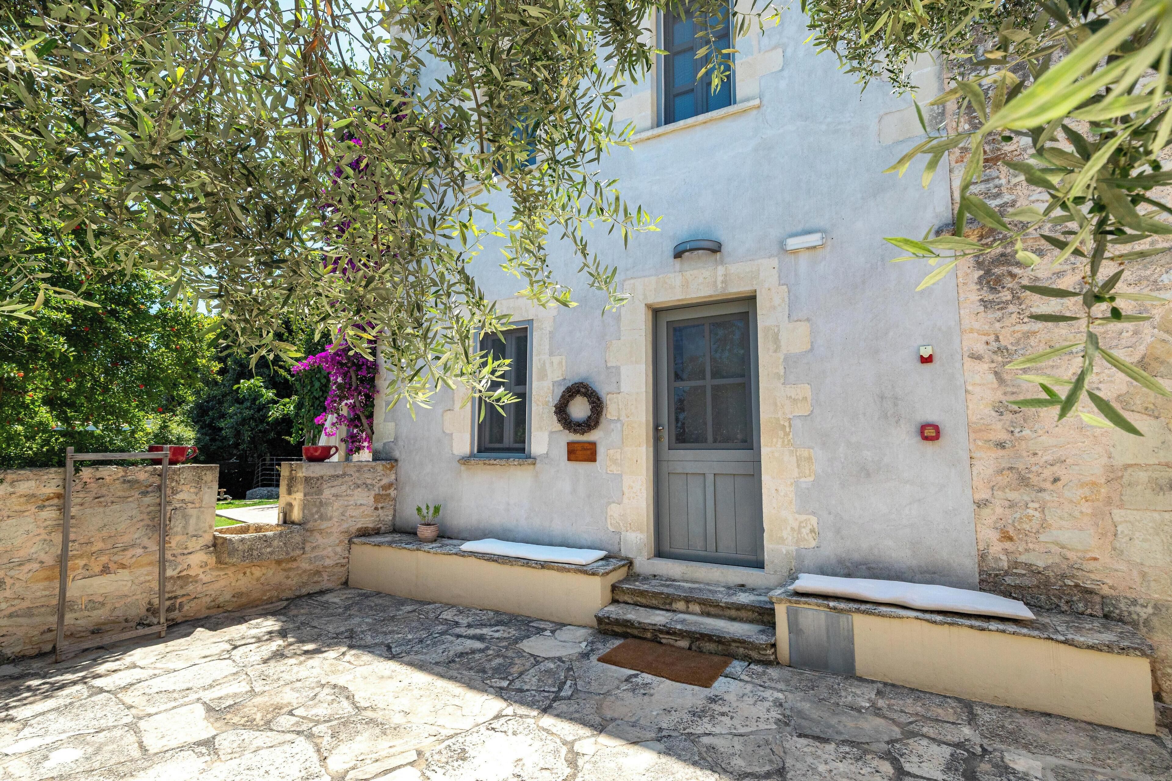 A stone-paved courtyard with a building featuring a grey door, windows, and a small bench.