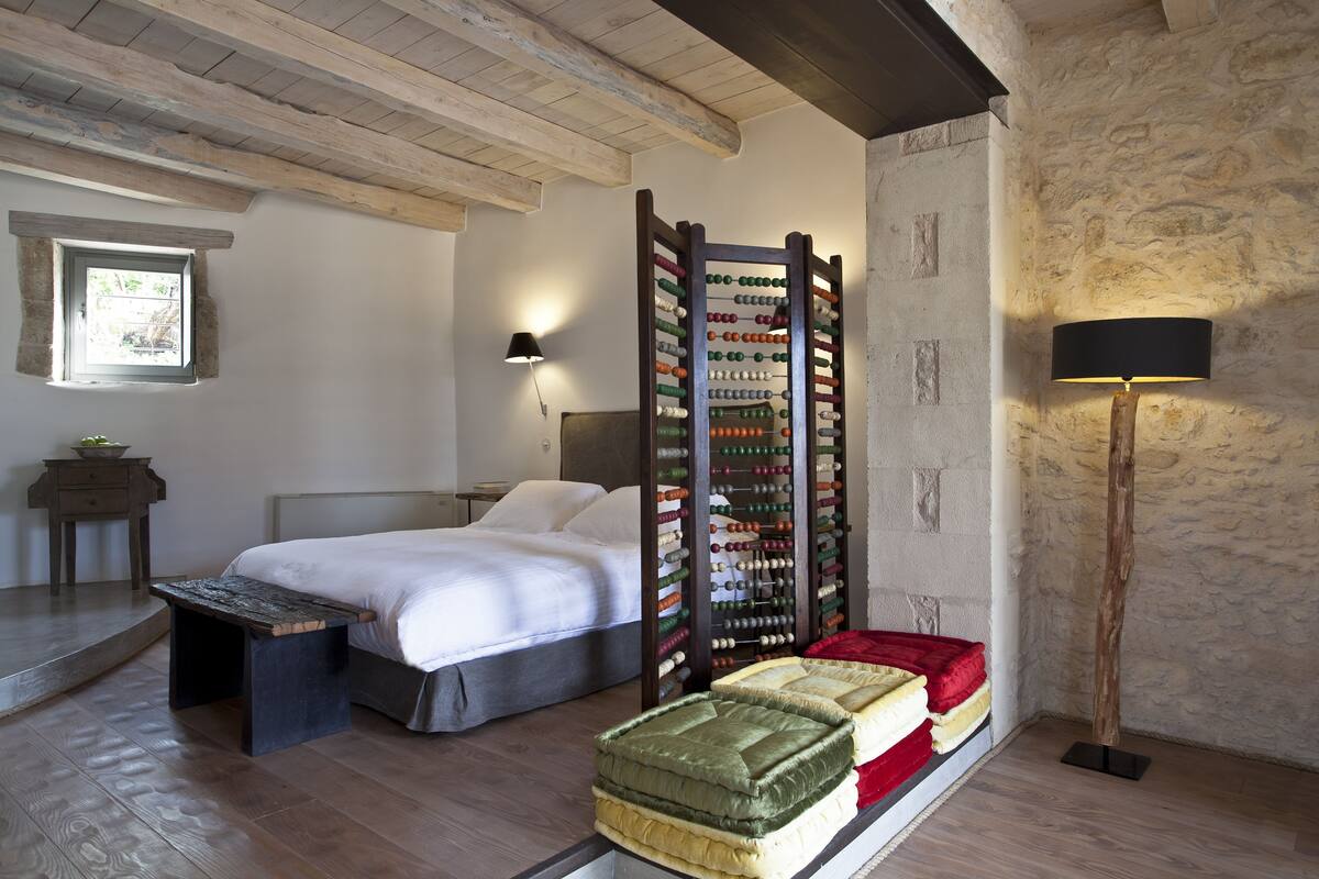 A bedroom with a bed, wooden headboard, a wall-mounted shelf with bottles, a bedside table, a lamp, and a window.