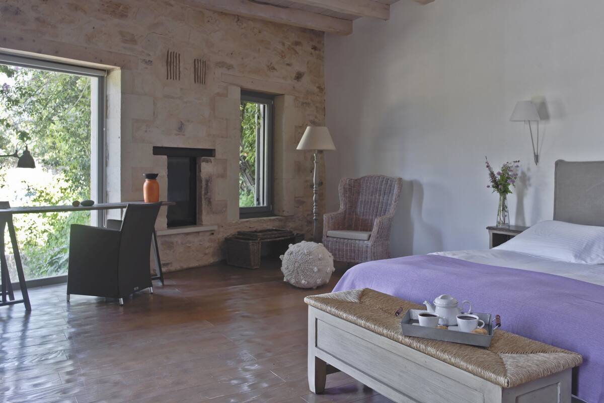 A room with a bed, a chair, a table, a lamp, and a fireplace.