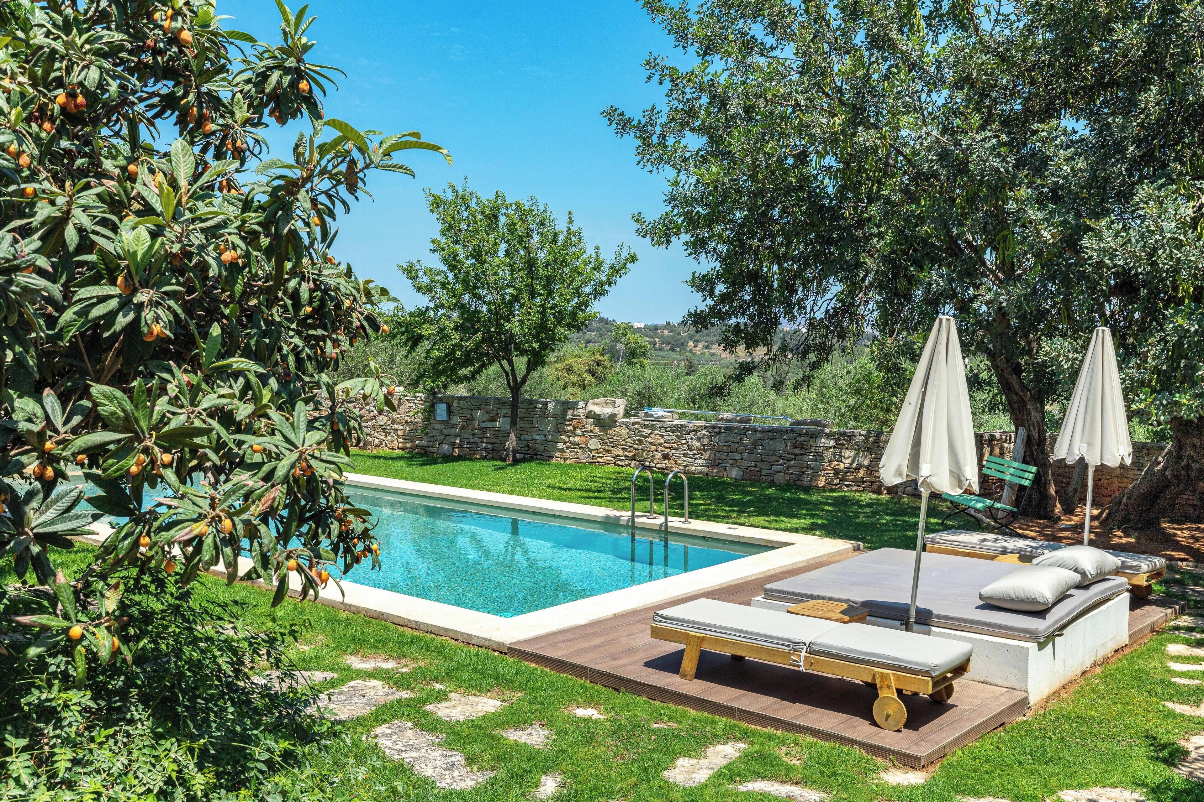 A swimming pool with lounge chairs and umbrellas, surrounded by greenery and stone walls.