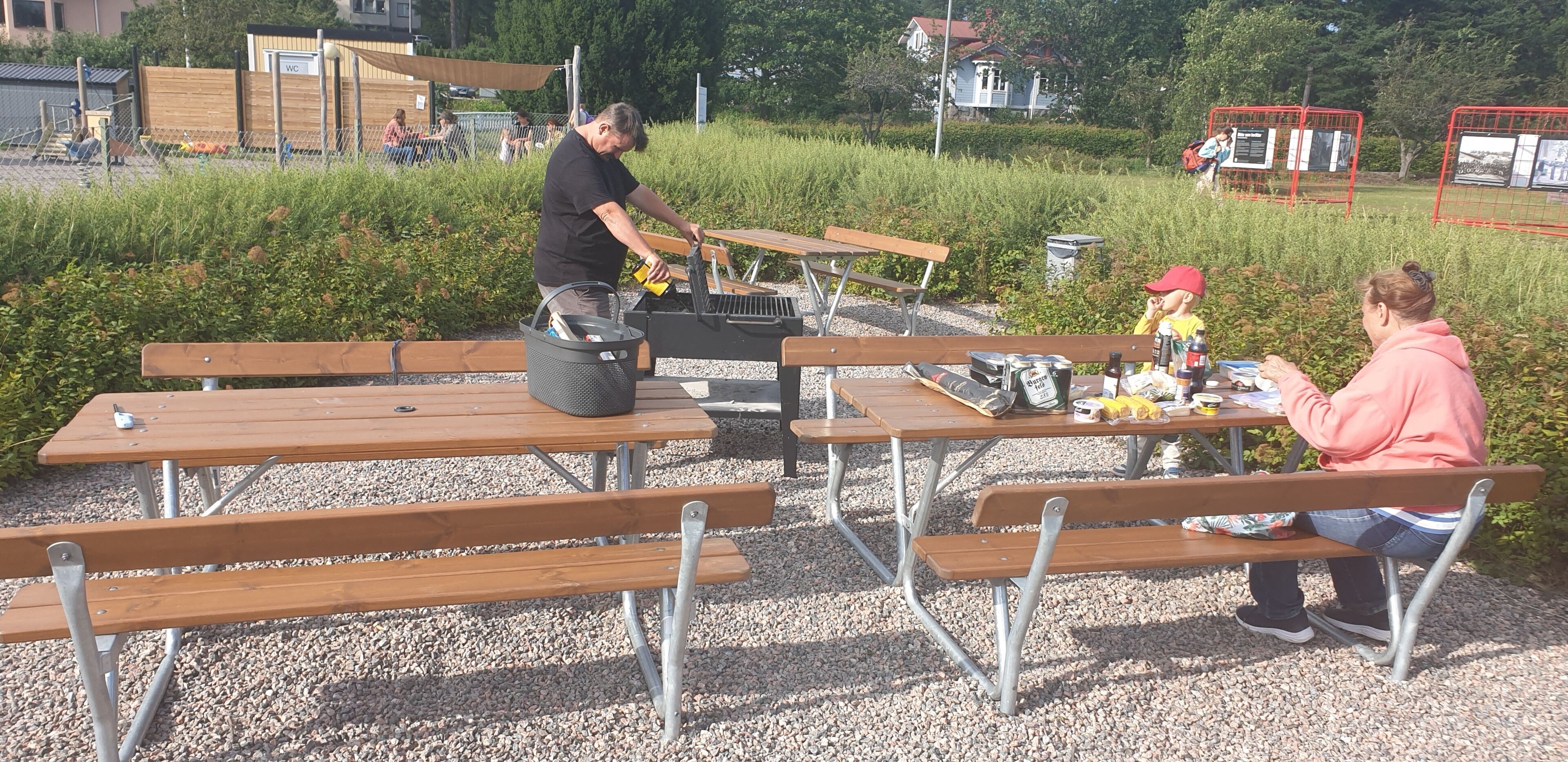 Picknicktische mit Bänken, ein Grill und ein Mülleimer im Freien.