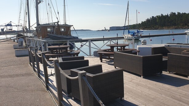 A wooden deck with wicker furniture, a small wooden structure, and a view of the water with boats. - Segelfartyget Libelle (Oxelosund)